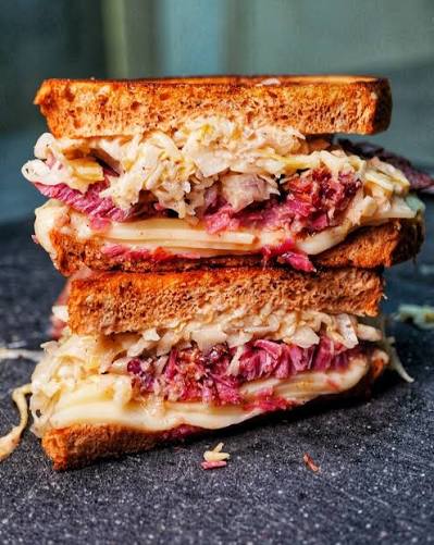 A close-up of a stacked grilled beef brisket sandwich on dark bread, filled with melted cheese, shredded red cabbage, and sauerkraut, set on a dark textured surface.