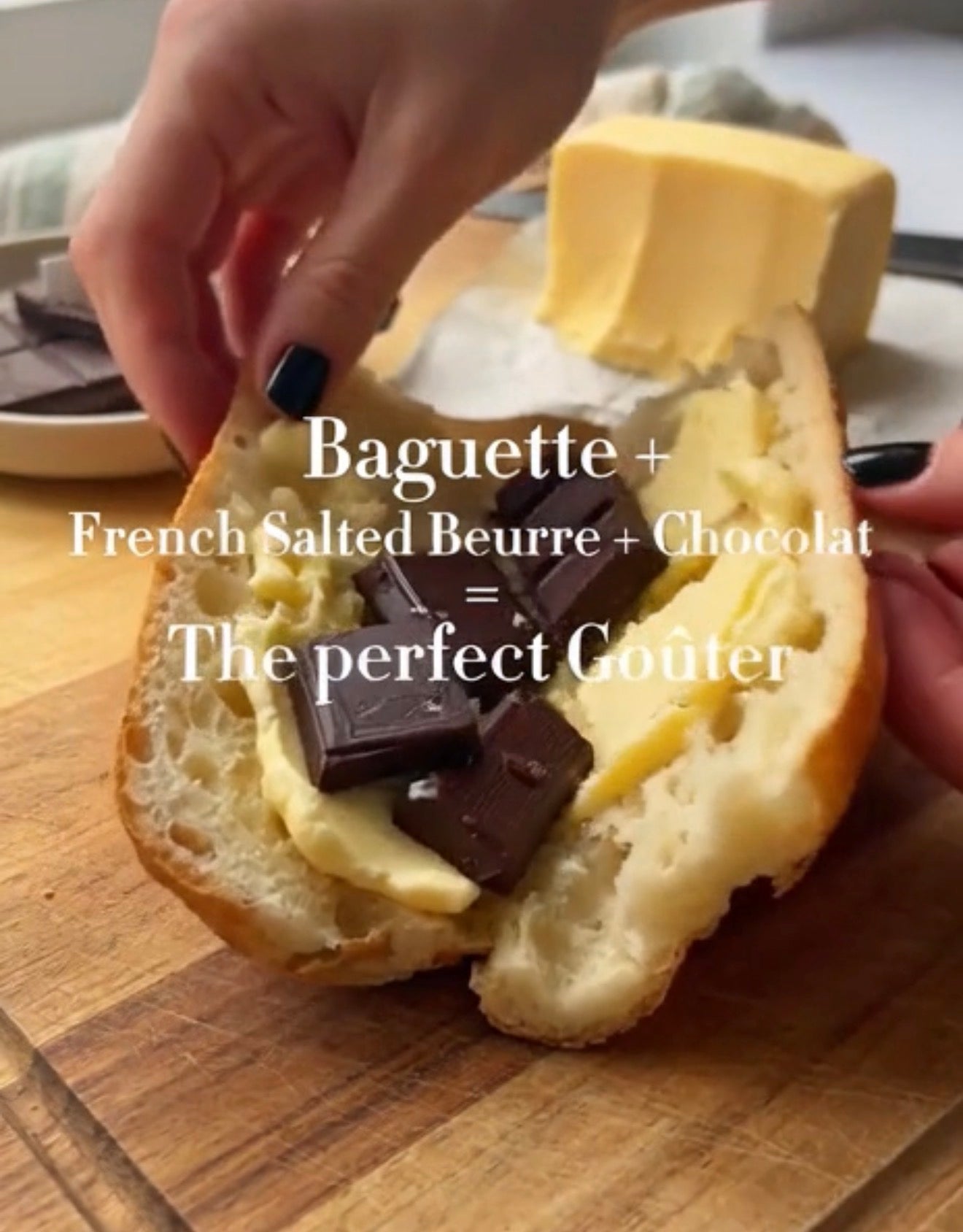 A close-up of a baguette being split open on a wooden cutting board, with butter and dark chocolate pieces being spread inside. A hand with black nail polish is visible holding the baguette, and another hand is at the right edge.