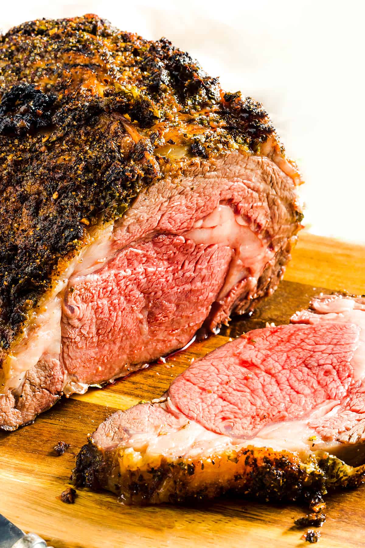 A close-up of a cooked beef prime rib roast with a dark, seasoned bark exterior and a pink, medium-rare interior, with a slice placed on a wooden cutting board.