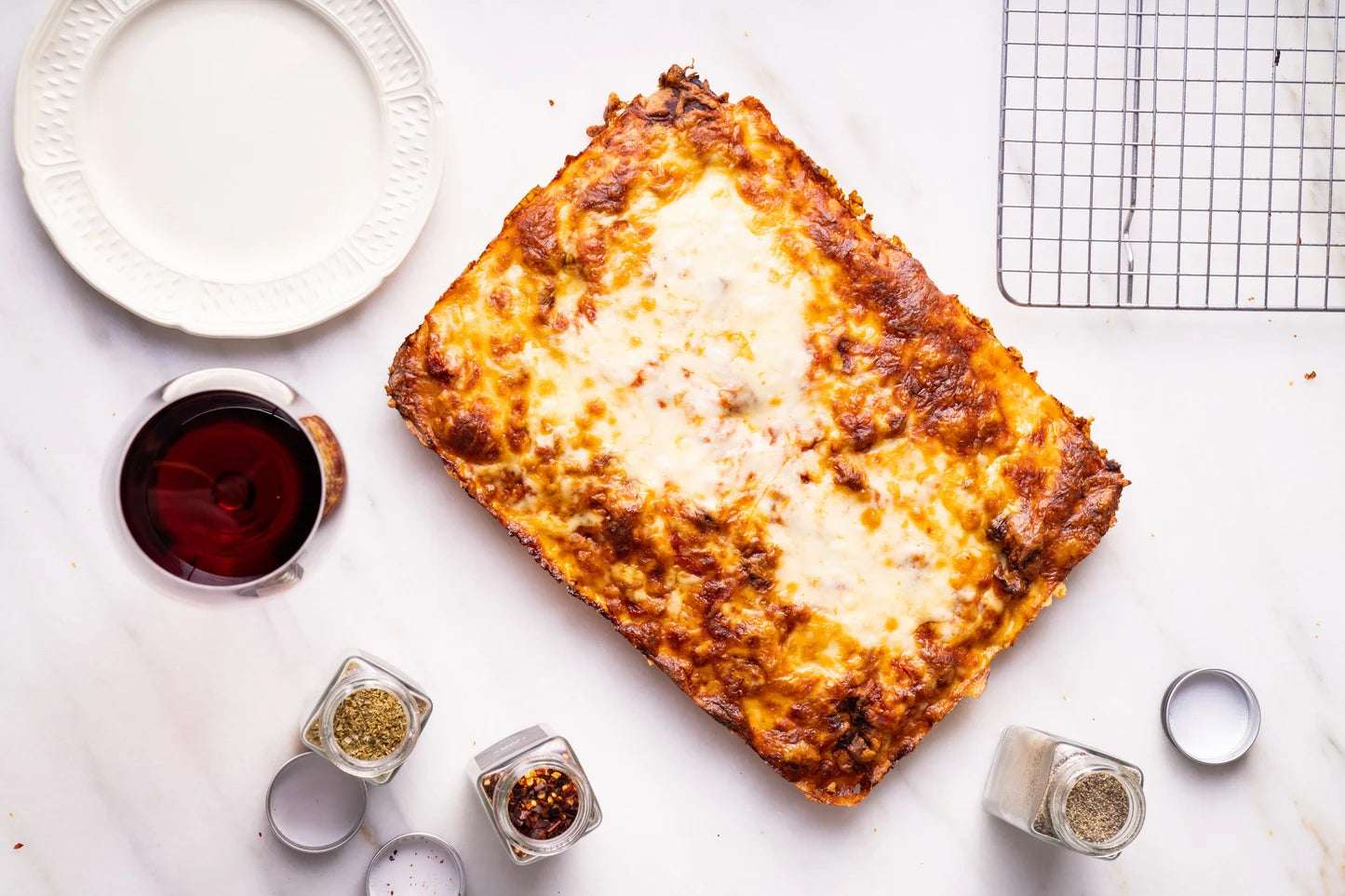 A baked rectangular casserole or lasagna with melted cheese on top, arranged on a white marble surface with a white plate, a glass of red wine, spice jars, and a cooling rack around it.