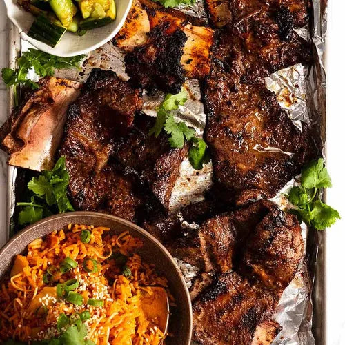 Grilled short ribs with charred marks on a foil-lined baking sheet, served with a side of orange rice and green vegetables.