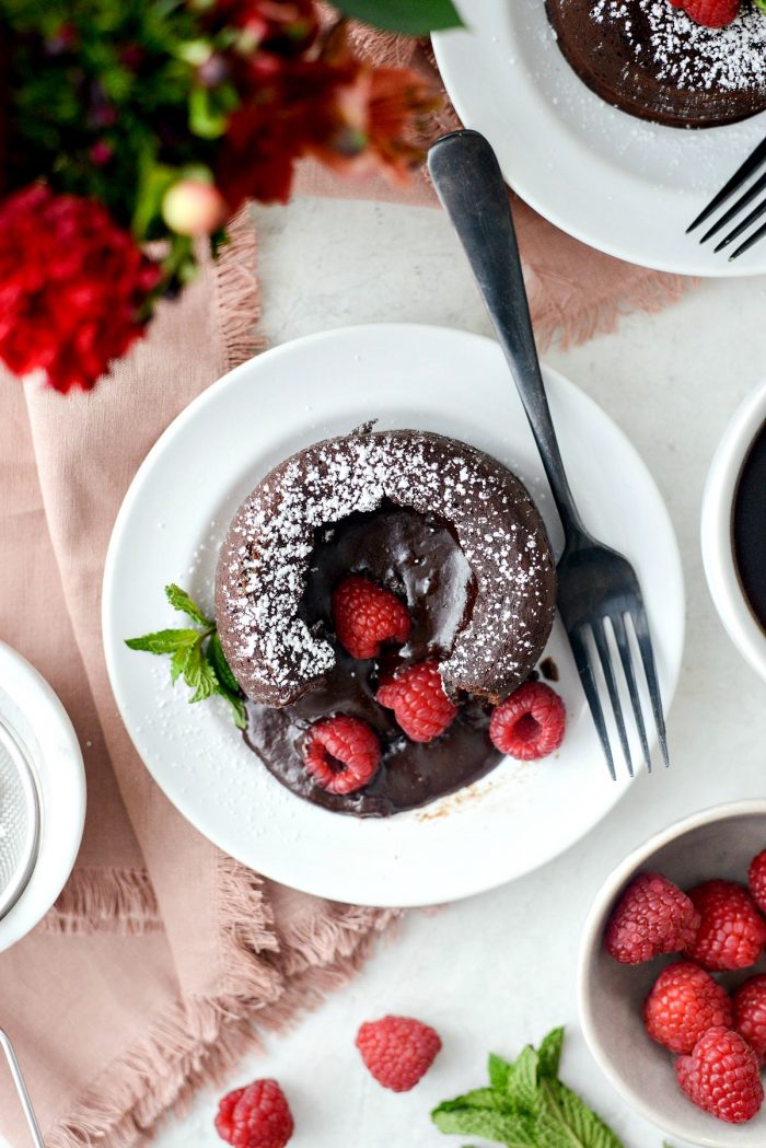 A chocolate lava cake on a white plate, topped with powdered sugar and fresh raspberries, with a fork on the side. The scene is styled with a pink napkin, red flowers, and bowls of raspberries.