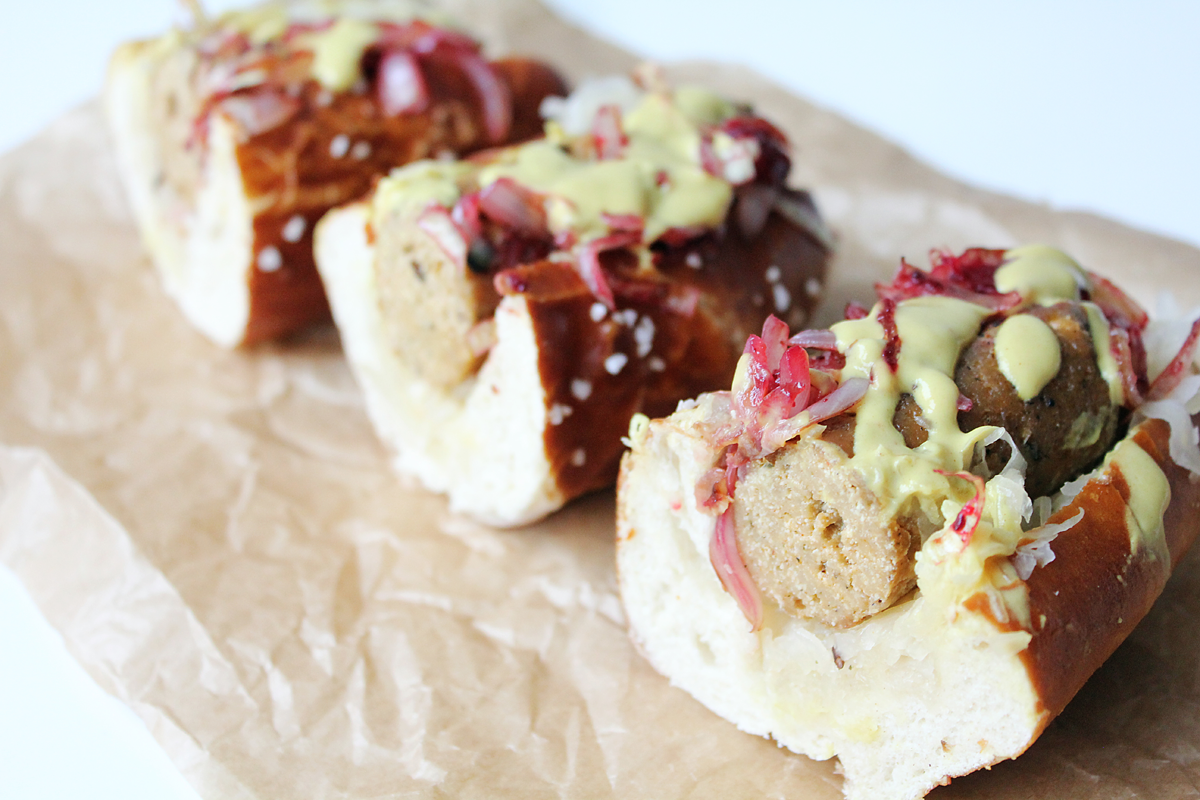 Three sausage-filled pretzel rolls topped with shredded cabbage, red onion and pale yellow sauce