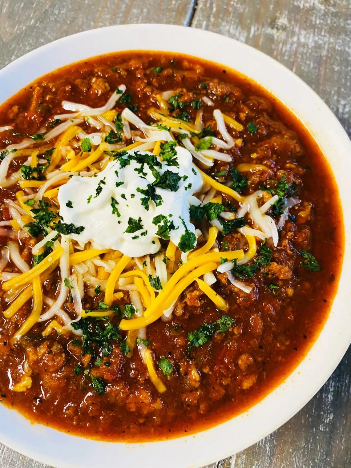 A top-down view of a bowl of chili ground chicken chorizo cheese soup with a white sour cream dollop, shredded yellow and cheddar cheese, and chopped green herbs on a rustic gray wooden table.