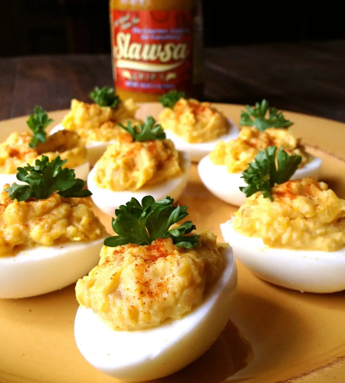 Deviled eggs on a yellow plate with a bottle of Slawsa sauce in the blurred background.