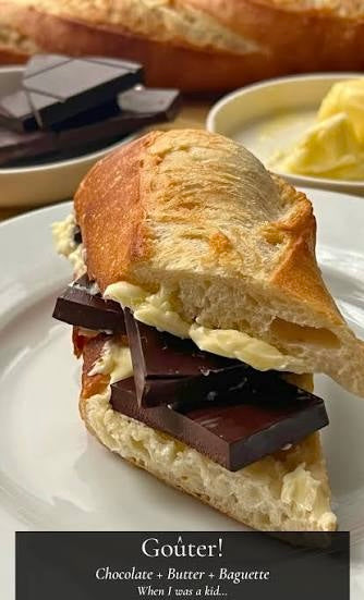 A close-up of a gourmet baguette sandwich filled with french butter and thick chocolate chunks, served on a white plate. In the background, there is a loaf of bread and a small bowl of butter.