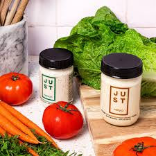 Two jars of Just Chipotle Ranch Dressing on a wooden cutting board with fresh tomatoes, carrots, and lettuce in a kitchen setting.