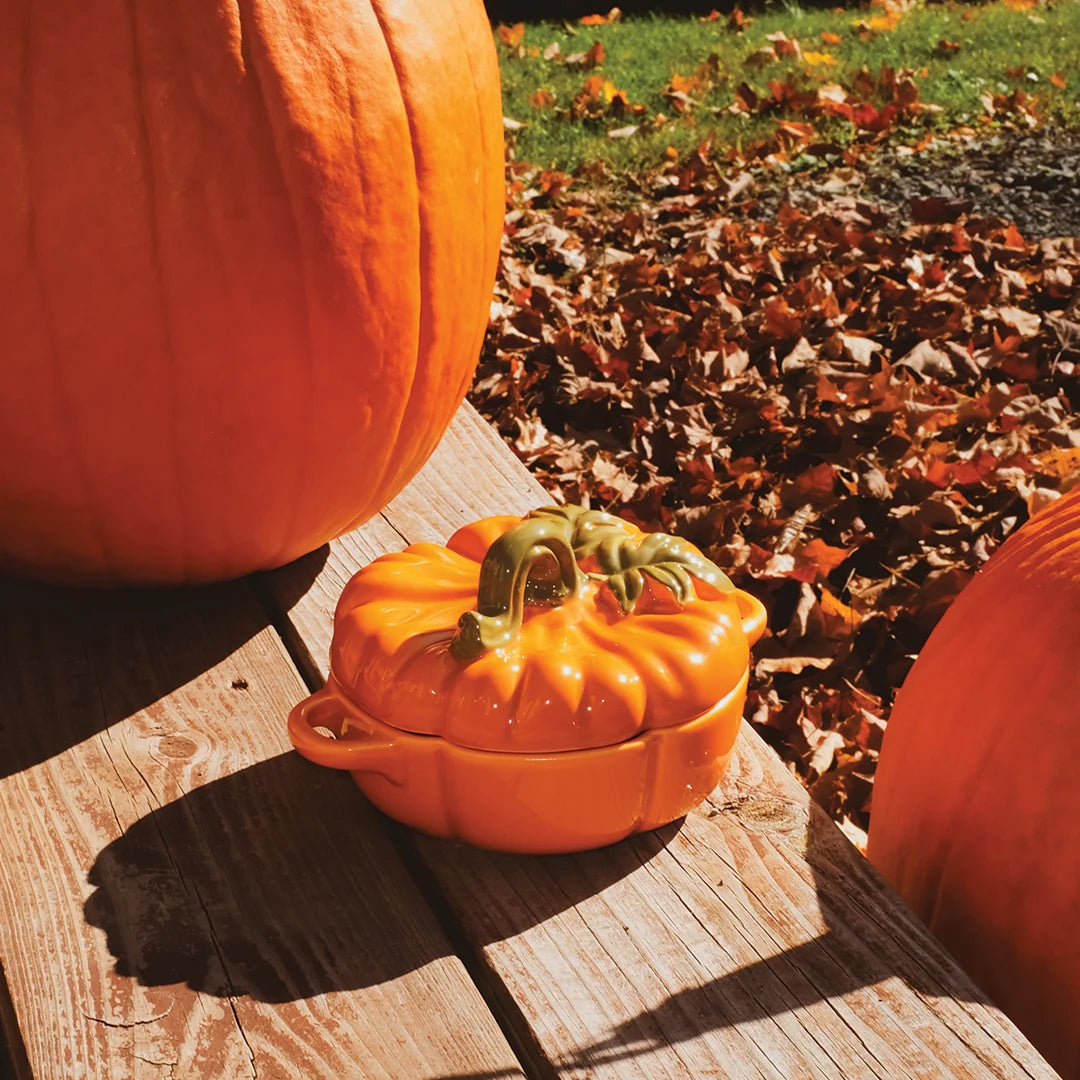 An orange pumpkin-shaped ceramic container with a lid and small side handles sits on a wooden table outdoors, with large pumpkins and fallen leaves in the background.