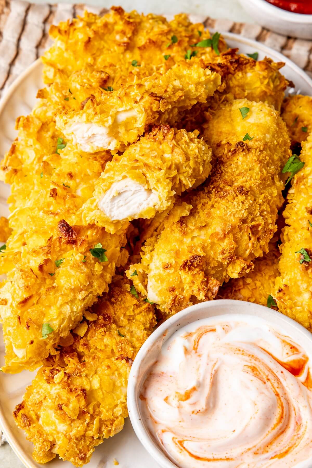 Crispy golden-brown fried chicken tenders on a white plate, garnished with chopped green herbs, served with a small white dipping sauce bowl.