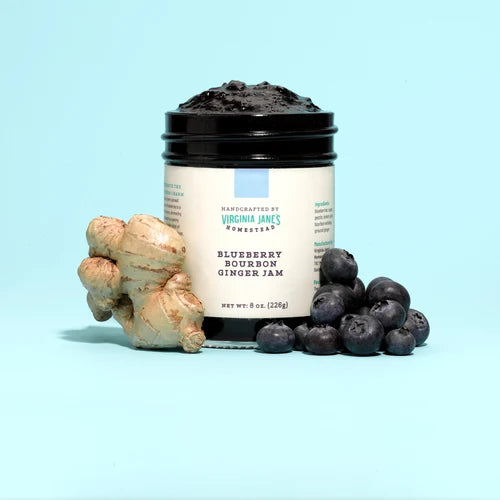 A jar of Virginia Jane's Handcrafted Bourbon Ginger Jam with blueberries and fresh ginger root props, set against a solid light blue background.