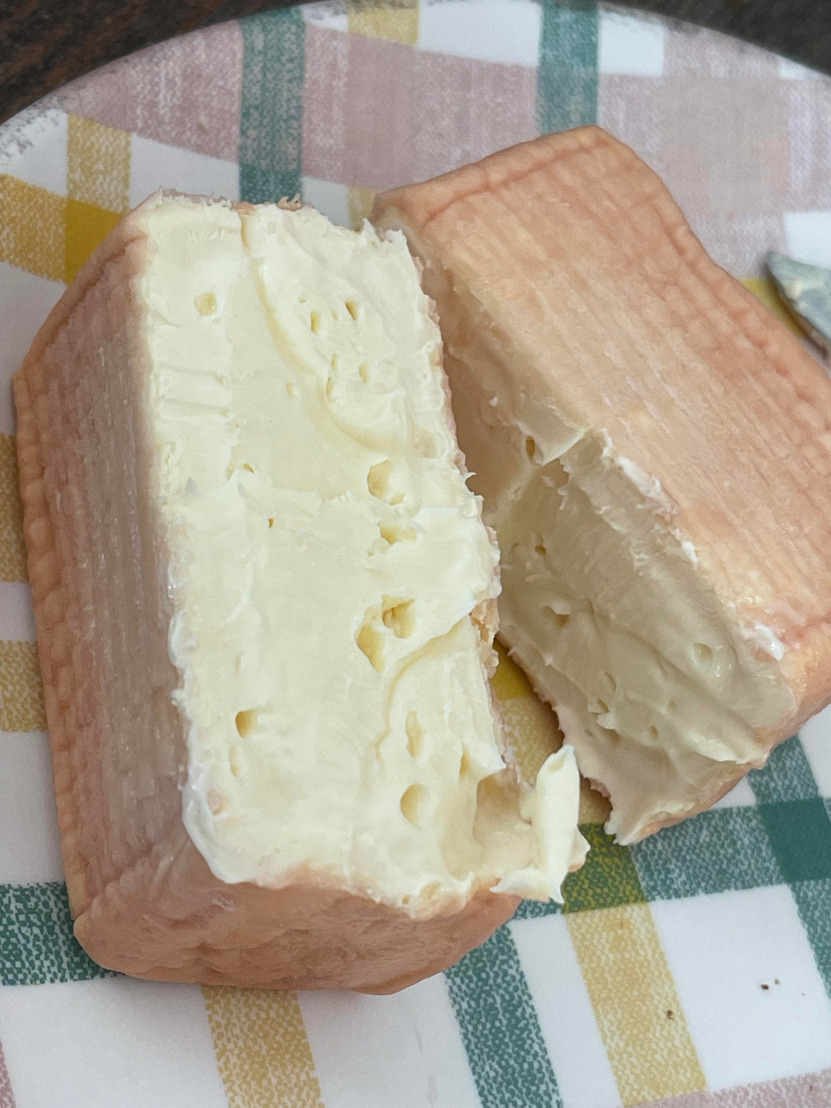 A close-up of Foxglove cheese cut into two pieces, showing a pale creamy off-white interior with small holes and a light tan rind, resting on a plaid tablecloth.
