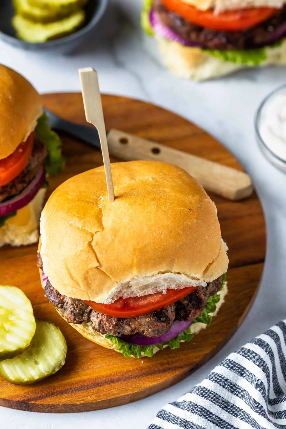 A close-up of a gourmet bison hamburger on a wooden board, featuring a golden-brown bun, beef patty, tomato, red onion, and lettuce, with pickles and a striped napkin nearby.