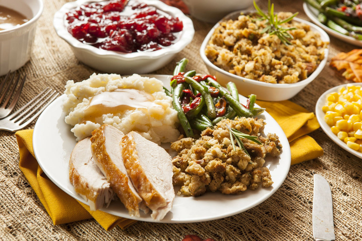 A Thanksgiving dinner spread on a burlap tablecloth, featuring a white plate with sliced turkey, mashed potatoes, stuffing, green beans, and cranberry sauce, surrounded by side dishes of corn and broccoli.