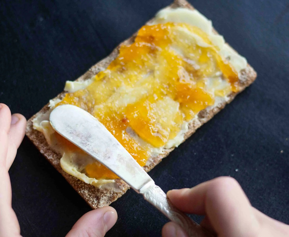A close-up of hands spreading orange fruit jam and cream on a slice of whole wheat bread using a silver butter knife, set against a dark slate background.