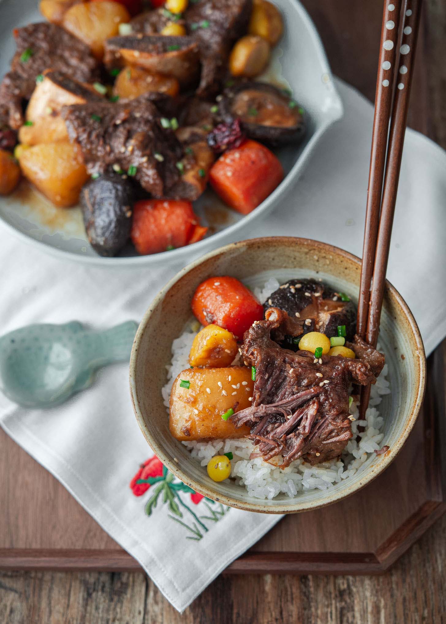 A bowl of beef stew with short ribs over white rice with carrots, potatoes, and mushrooms, served with chopsticks, on a wooden tray with a spoon and a larger plate of the same stew in the background.