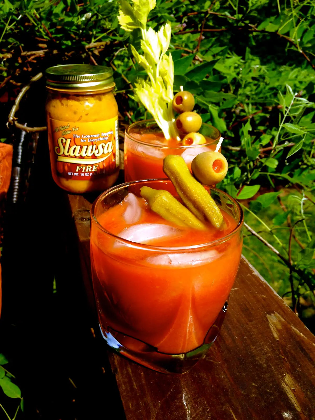 A glass of red Bloody Mary cocktail garnished with pickles, olives, and a celery stalk, with a jar of Slawssa Fire mustard sauce standing behind it on a wooden surface outdoors.