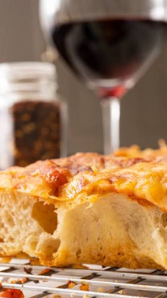 A close-up of a slice of cheesy bread or focaccia on a cooling rack, with a blurred glass of red wine and a jar in the background.