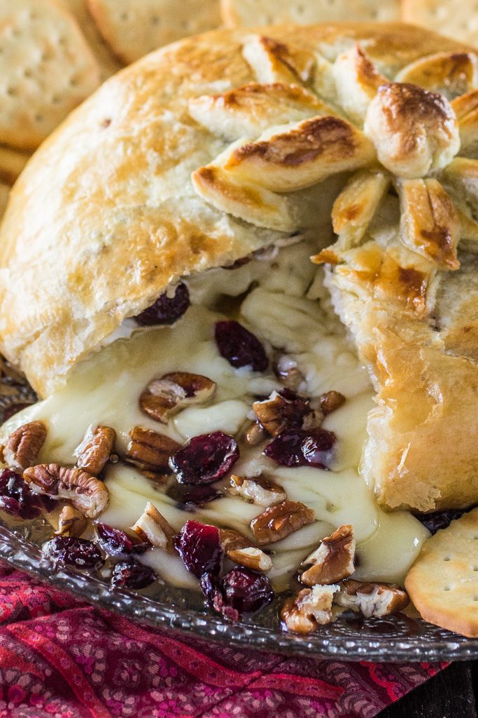 A close-up of a baked cheese ball appetizer with a golden flaky crust, melted cheese, pecans, and dried cranberries, served on a dark plate with crackers.