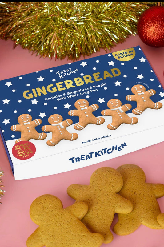 A box of Treat Kitchen Gingerbread cookies with several gingerbread people cookies placed in front on a pink surface, with gold tinsel and a red ornament in the background.