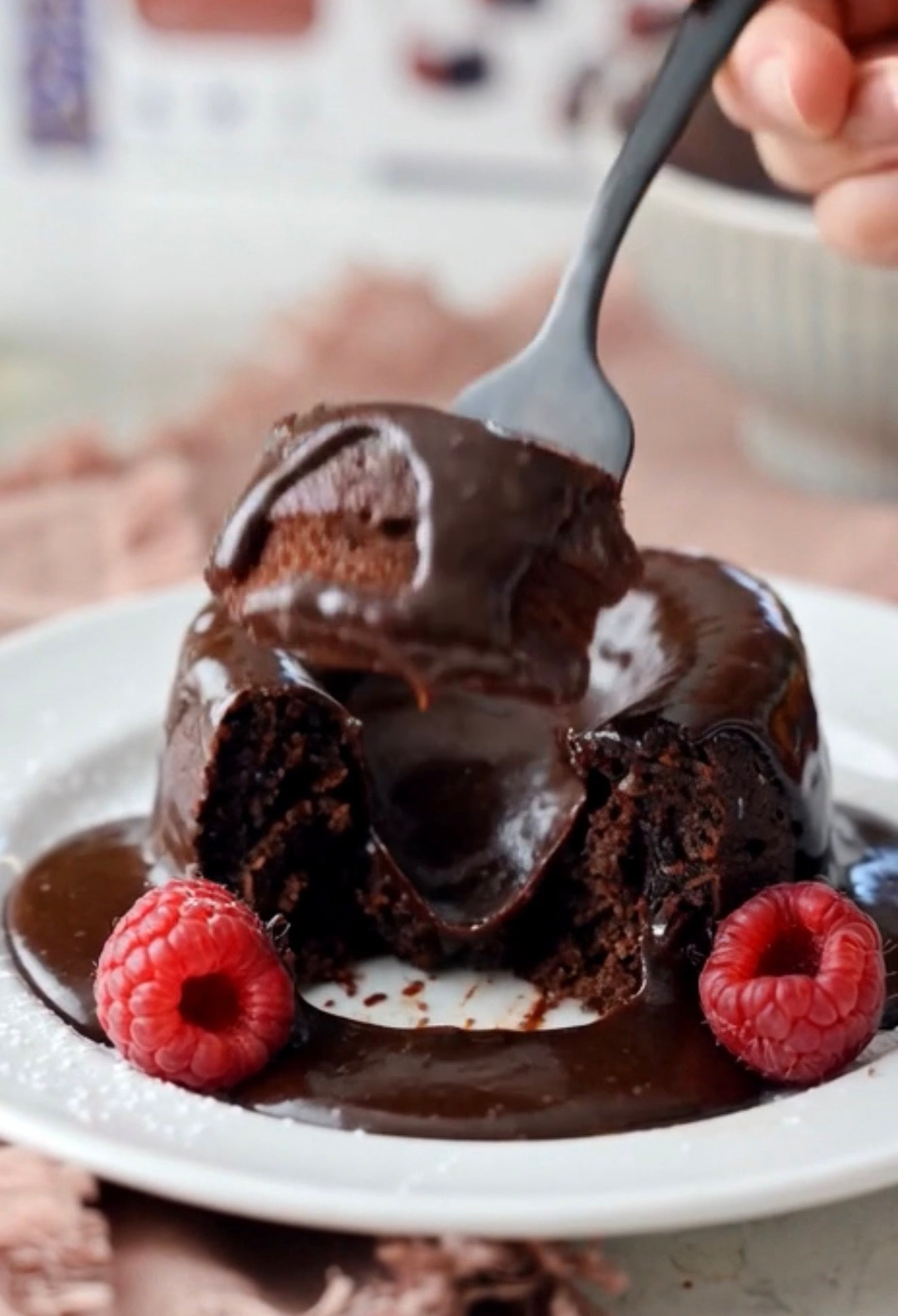A close-up of a chocolate lava cake on a white plate, drizzled with dark chocolate sauce and garnished with two fresh raspberries. A hand holds a fork lifting a piece of the cake, with a blurred kitchen or dining background.