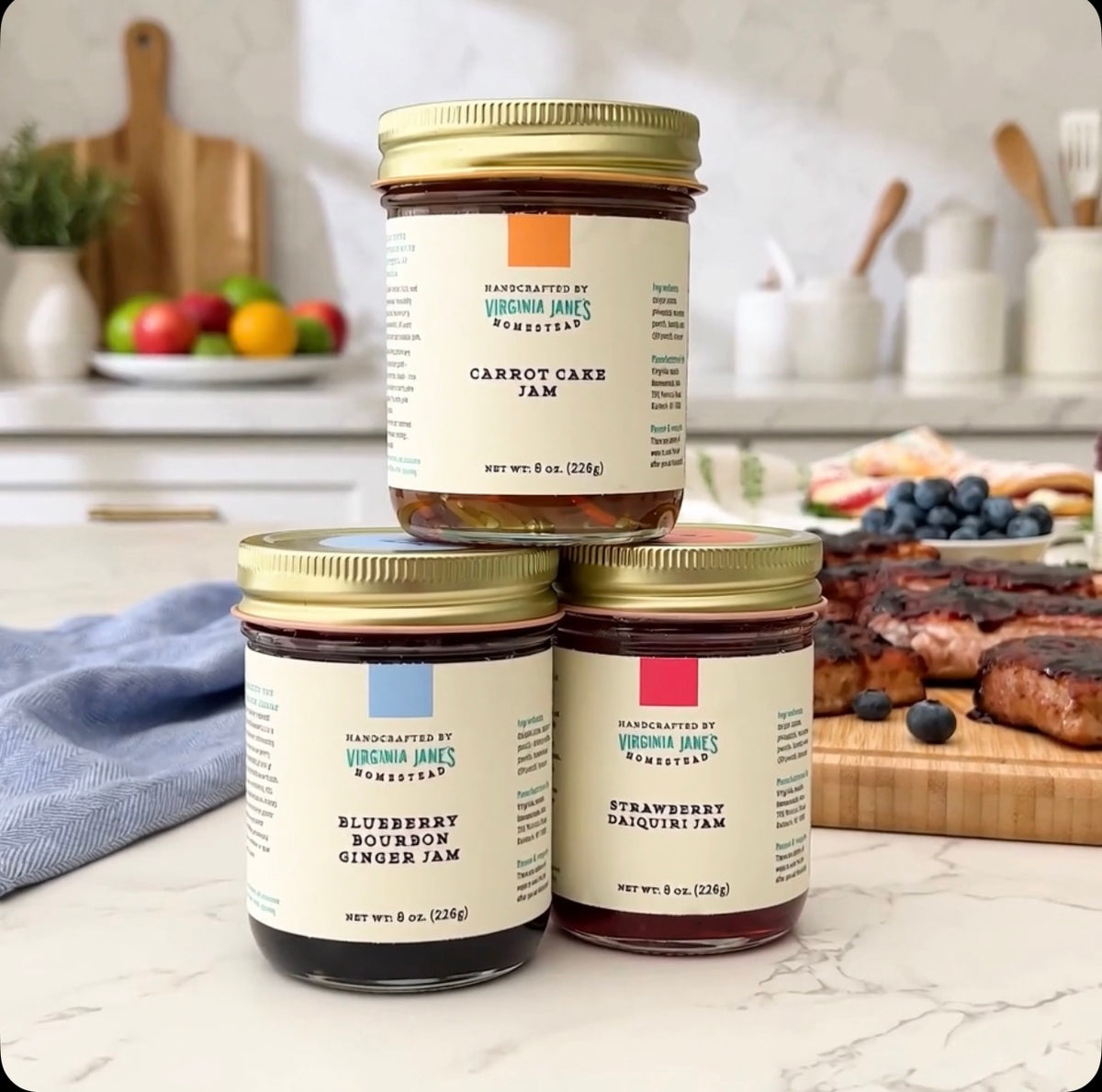 Three jars of Virginia Jane's Handmade Jam arranged on a marble countertop in a kitchen setting, with blueberry pancakes and cooked meat visible in the background.