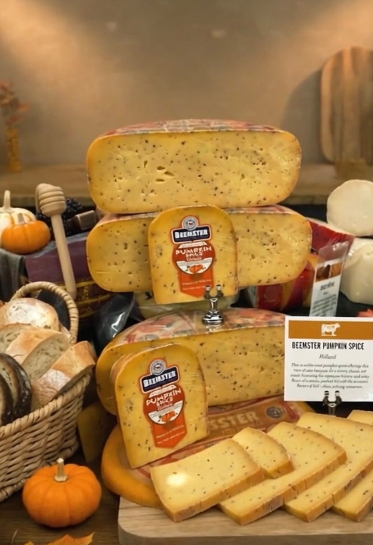 A display of Beemster cheese products, including stacked whole and sliced wheels of cheese with a small sign reading 'Beemster Pumpkin Spice,' set on a table with bread, pumpkins, and warm autumn decor in the background.