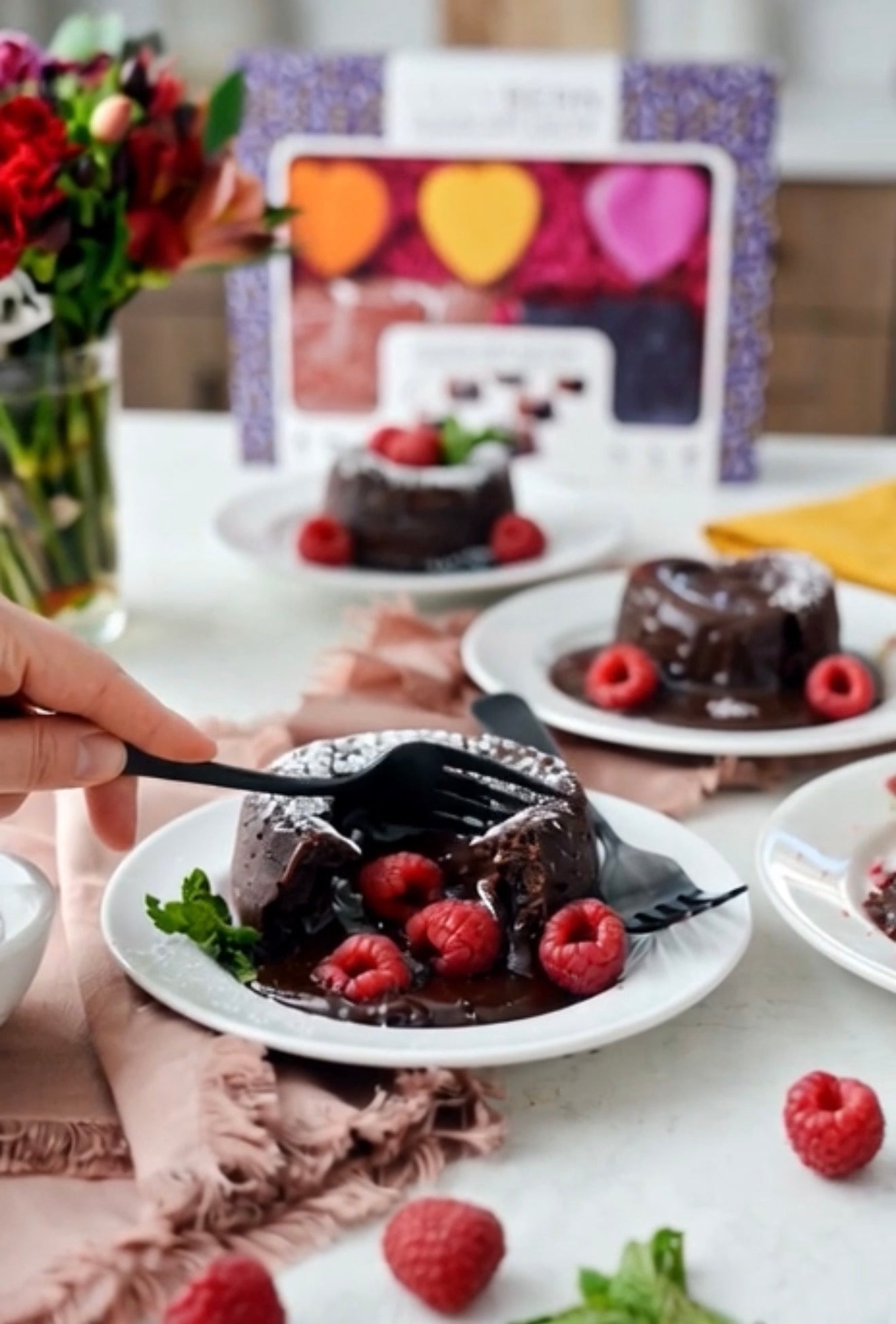 A chocolate lava cake dessert with raspberries on a white plate, with a hand holding a black spoon and fork. Another plate with a whole cake and a decorative heart-patterned box are visible in the background on a table with flowers and a pink napkin.