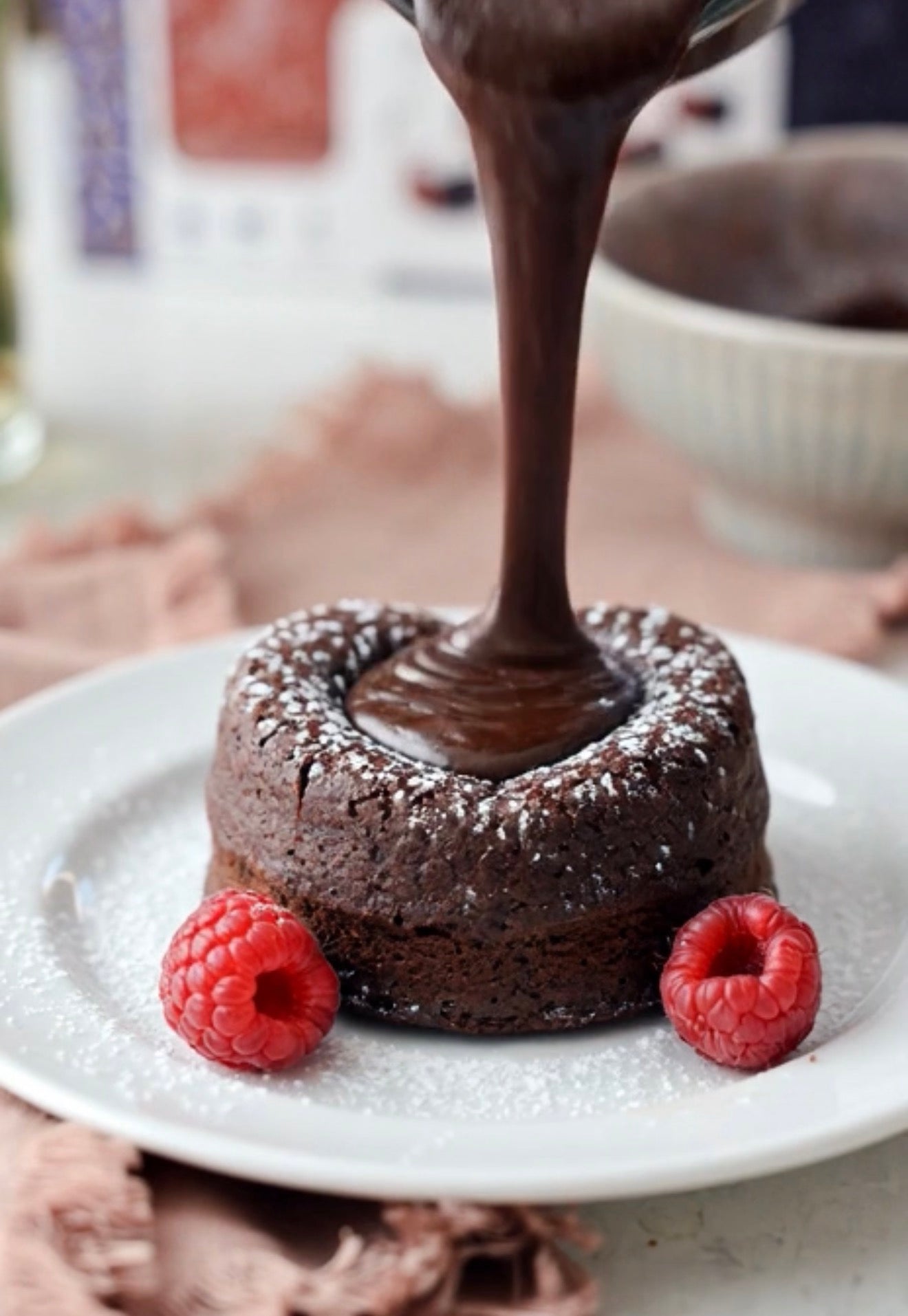 A small dark chocolate lava cake on a white plate, with thick melted chocolate being poured into the center and two fresh raspberries on either side.