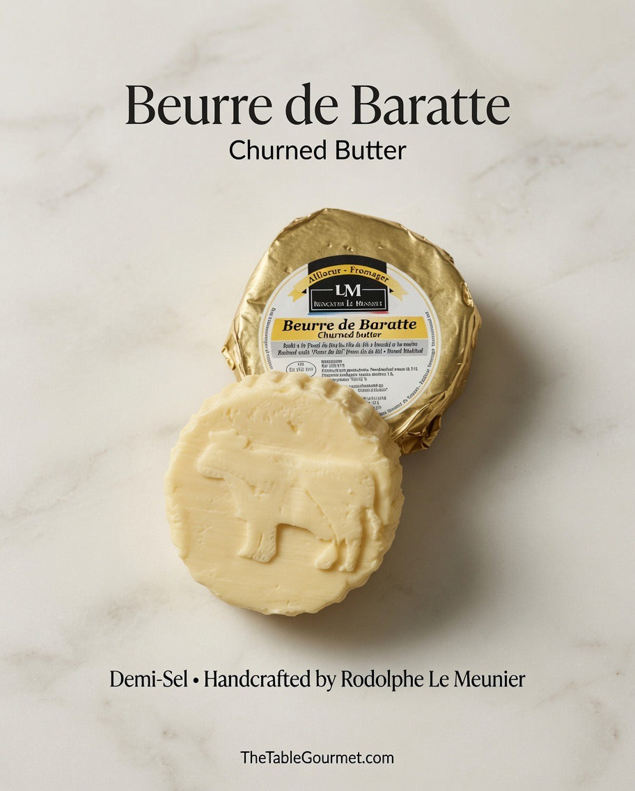 A round bar of churned butter in front of its gold foil wrapper on a light marble surface, with text reading 'Beurre de Baratte' and 'Churned Butter' plus 'TheTableGourmet.com'.