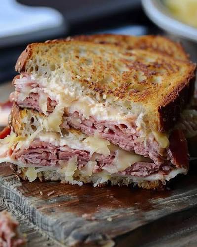 A close-up of a toasted Reuben sandwich on a wooden board, showing layers of sauerkraut, sliced deli meat, melted cheese, and melted butter.