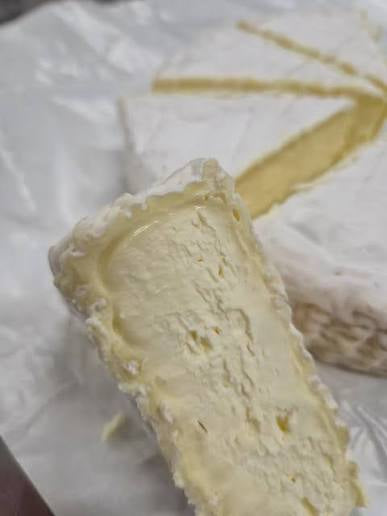 A close-up of a wedge of soft white cheese, likely brie or camembert, with a pale yellow interior, resting on white waxed paper with another slice blurred in the background.