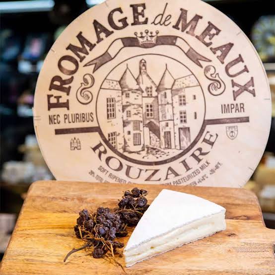 A wedge of Rouzaire Fromage de Meaux soft cheese on a wooden board with dried grapes, in front of a large branded wooden sign.