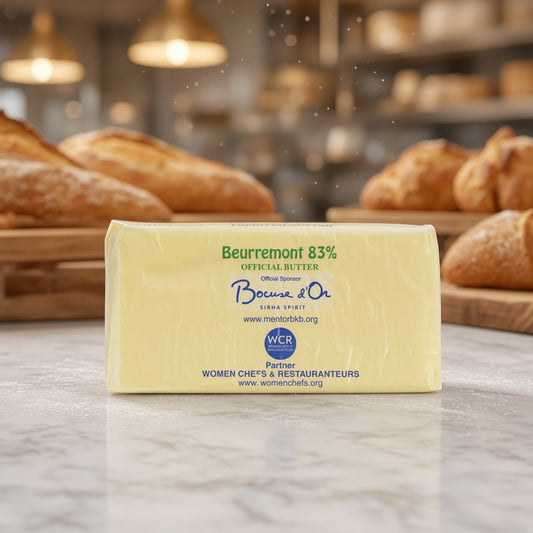 A stick of Boucmont & O. Beurremont 83% Official Butter centered on a light marble countertop, with a blurred bakery background showing bread loaves and warm hanging lights.