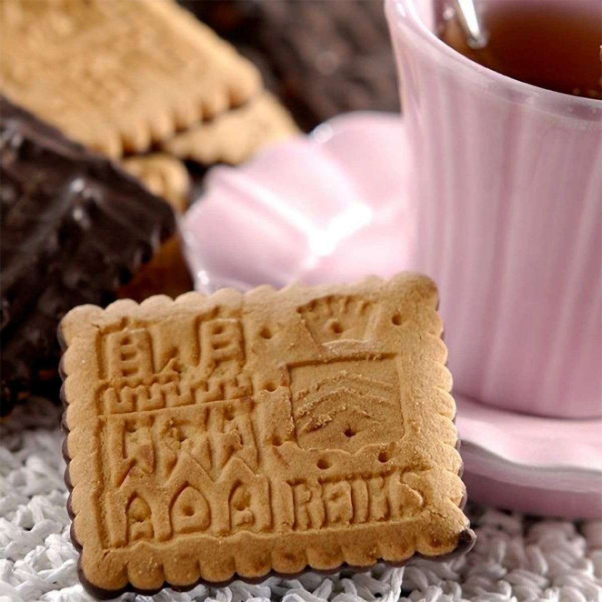 A square golden-brown biscuit with embossed decorative lettering and border, styled on a white textured surface next to a pink cup of tea and chocolate pieces.