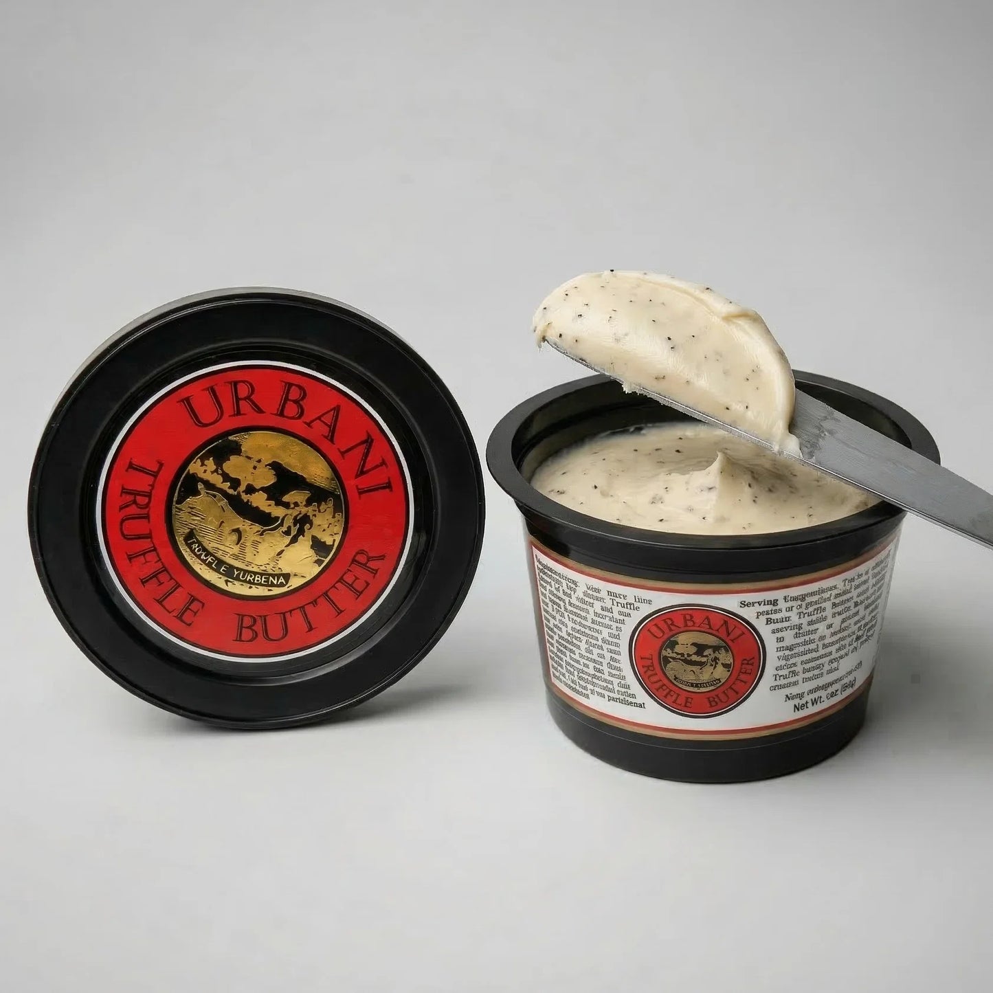 Urbani Artisan Truffle Butter in a black plastic tub, shown open with a knife lifting a scoop of creamy beige truffle butter. The red and black lid is placed beside the tub on a light gray background.