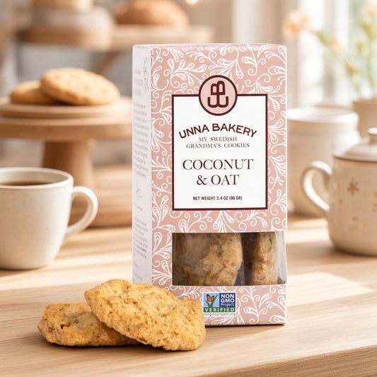 A box of Unna Bakery Coconut & Oat cookies sits on a wooden table with two cookies in front and cups in the softly blurred background.