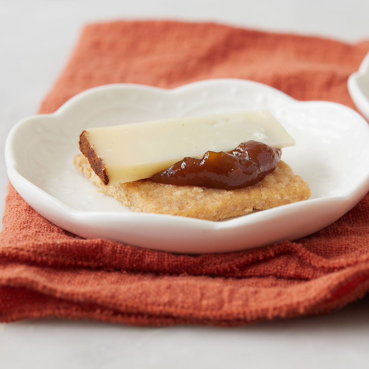 A close-up of a small white scalloped dish holding a cracker topped with a slice of white cheese and a dollop of dark brown spread, set on an orange textured cloth.