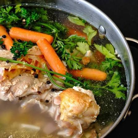 A close-up of a rustic chicken and vegetable soup in a silver metal pot, featuring baby carrots, fresh green parsley, and chives floating in a clear broth.