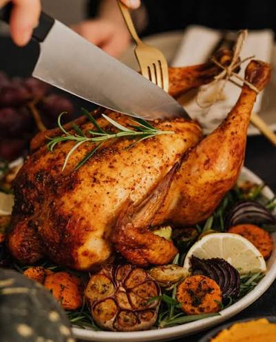 A roasted whole chicken on a white platter with roasted root vegetables, rosemary sprigs, a lemon wedge, and a knife being used to cut into the chicken. A hand is visible holding the knife.