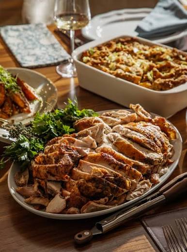 A plated sliced roasted turkey on a dining table, with a side of greens, a casserole dish, a wine glass, and table setting visible in a warm indoor dining environment.