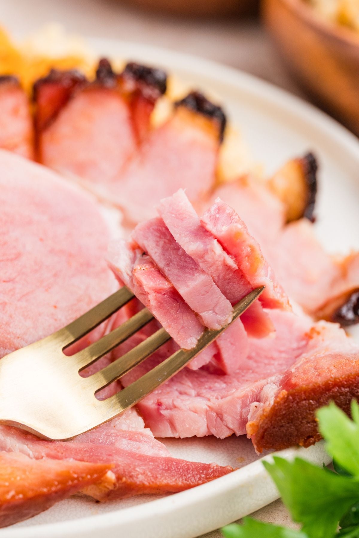 Sliced pink ham or roast on a white plate, with a gold fork lifting a piece in the foreground and blurred garnish in the background.