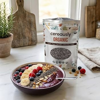 A bowl of purple smoothie topped with Chia seeds banana slices, berries, and granola sits on a kitchen counter next to a stand-up pouch of cereausly Organic chia seeks. The scene is styled with a wooden cutting board, a potted plant, and a folded cloth in a bright kitchen setting.