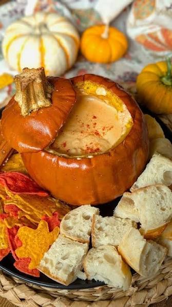Pumpkin soup served in a carved orange pumpkin bowl, surrounded by bread cubes, fall leaves, and small pumpkins on a woven tray.