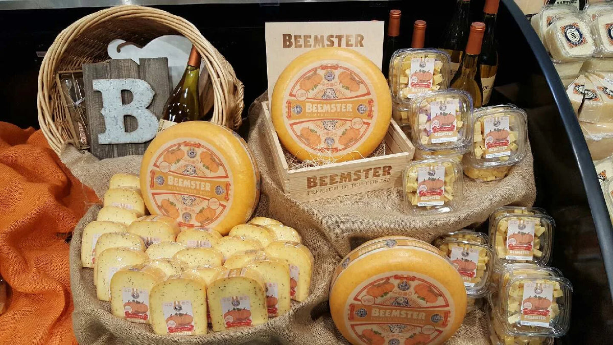 A display of Beemster cheese products, including whole round cheeses, sliced cheese wedges, and packaged cheese cubes, arranged on burlap with a wicker basket and wooden sign in the background.