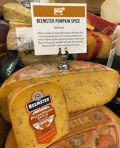 A wheel of Beemster Pumpkin Spice Gouda cheese displayed in a retail setting, with a white sign above reading 'Beemster Pumpkin Spice Holland' and a product label in the foreground.