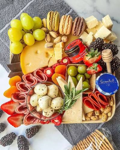 A beautifully arranged charcuterie board featuring an assortment of cheeses, crackers, fruits, cured meats, nuts, and sweets on a gray textured cloth over a white marble surface.