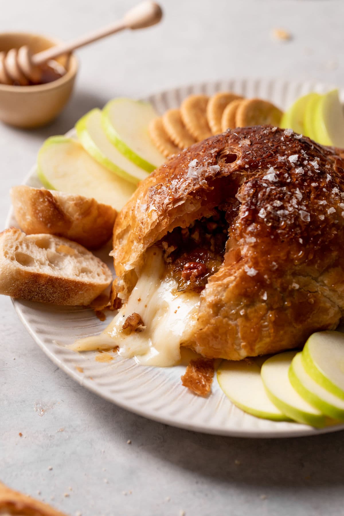 A close-up of a baked pastry filled with melted cheese and topped with coarse salt, served on a white plate with sliced green apples, crackers, and a small bowl of honey dipper in the background.