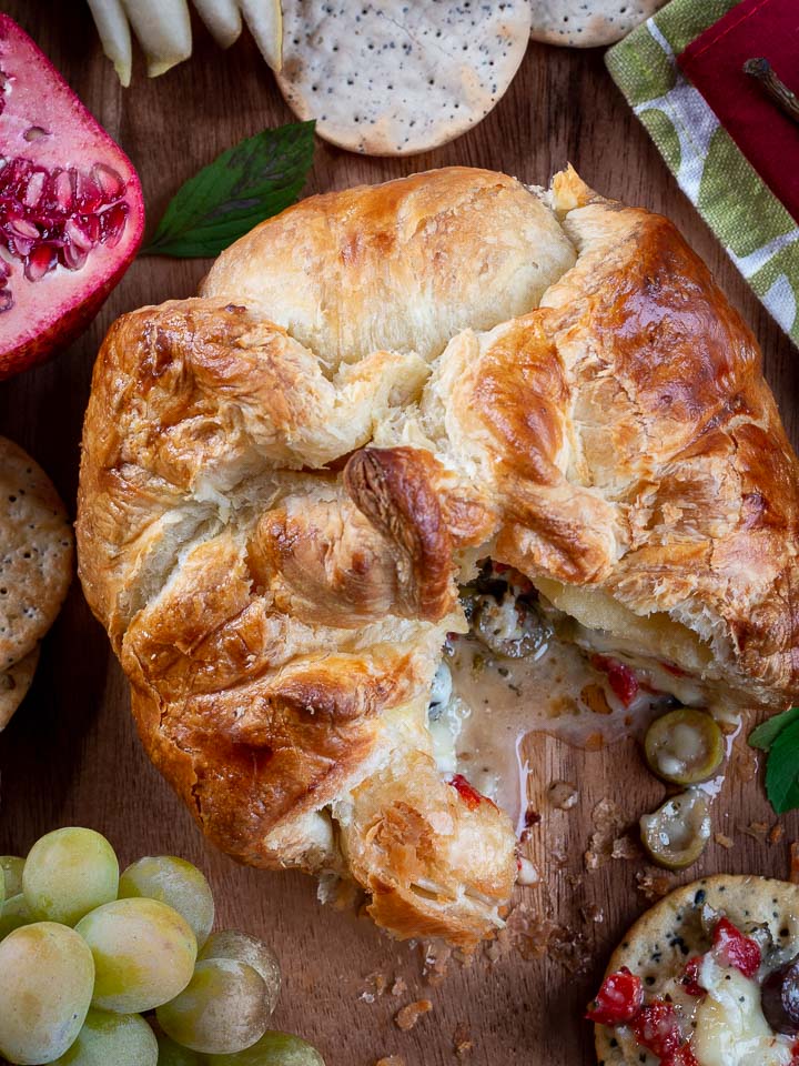 A golden-brown flaky pastry bread, cut open to reveal a savory filling with mushrooms, olives, and red peppers, served on a wooden board with crackers, grapes, a pomegranate, and a green cloth napkin.