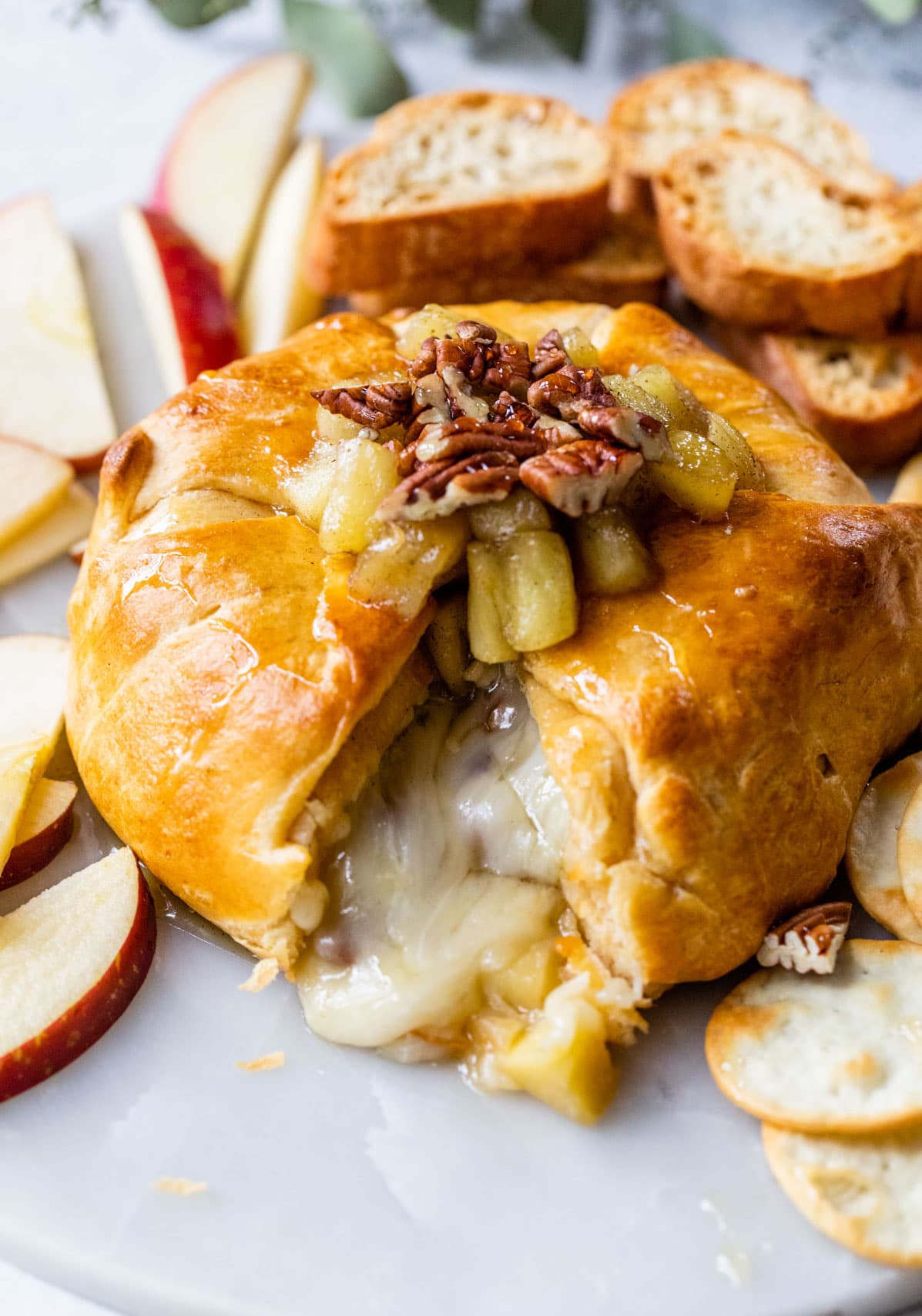 A baked cheese pastry with a golden-brown crust, topped with apple pieces and pecans, with melted cheese oozing out. Sliced apples, bread, and crackers are arranged around it on a white plate.