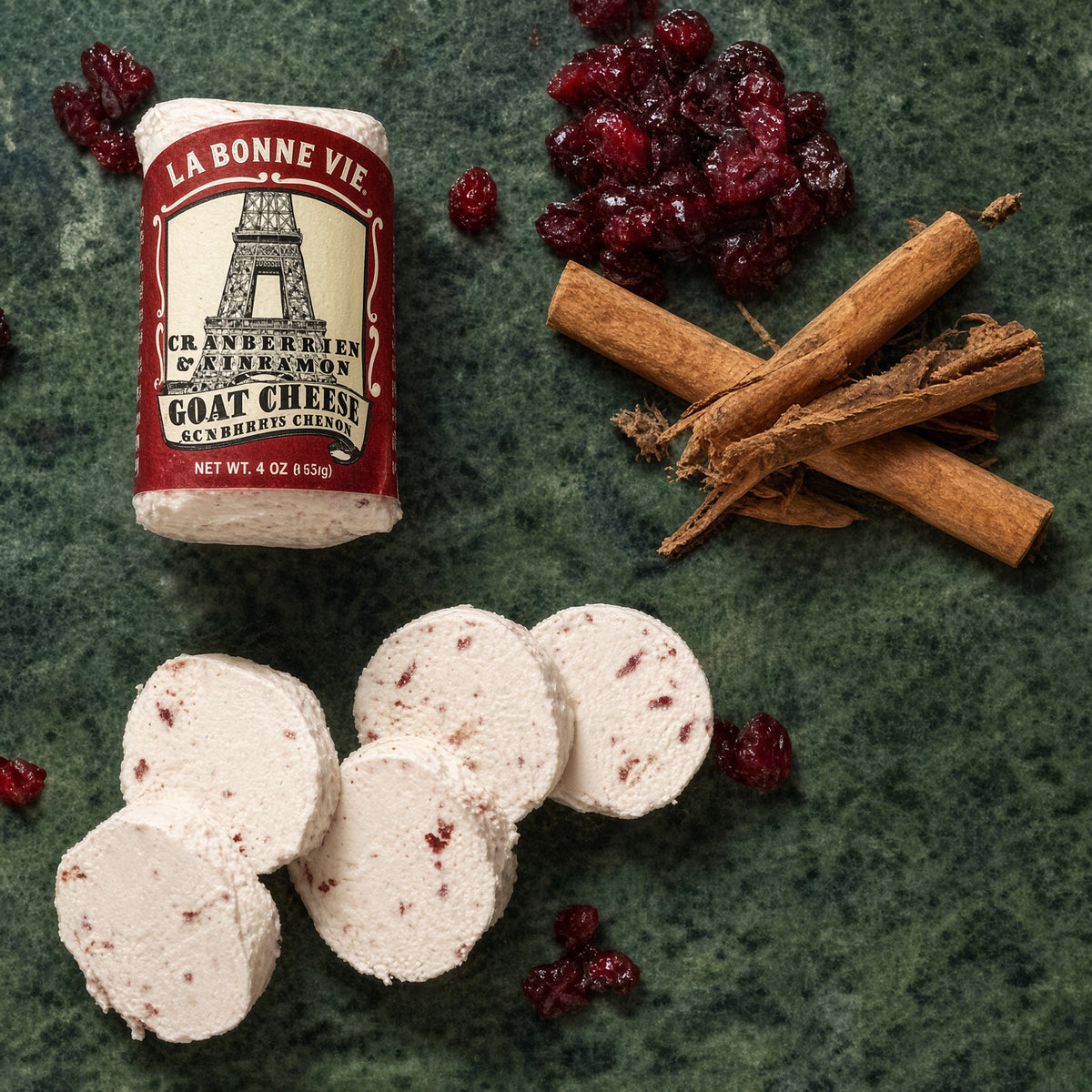 A 4 oz wheel of La Bonne Vie Cranberry Cream Cheese with several sliced rounds arranged on a dark green textured surface, alongside dried cranberries, cinnamon sticks, and a few whole cheese wheels. The product label is visible, and no person or real environment is present, so this is Product/Packaging rather than Lifestyle.